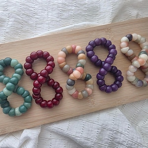 Può includere: Cinque elastici per capelli circolari disposti in fila, ciascuno composto da una serie di piccole perline sferiche lisce. I colori includono verde acqua scuro, bordeaux intenso, un mix di pesca e grigio, viola scuro e una miscela di colori chiari.