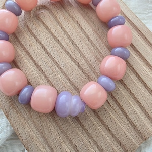 May include: A stretch bracelet featuring alternating smooth, rounded peach-colored beads and smaller, glossy lavender-colored spacer beads. The beads are arranged in a circular pattern, creating a simple, uniform design with a polished finish.