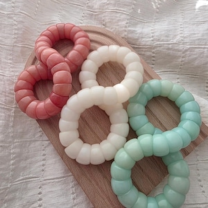 Puede incluir: Cuatro anillos de cuentas de colores en tonos rosa, blanco y verde azulado están dispuestos sobre una pequeña tabla de cortar de madera. Los anillos tienen un diseño texturizado y segmentado. El fondo es una tela blanca texturizada.