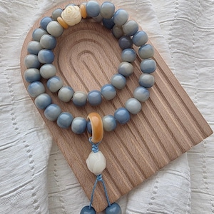 May include: A beaded bracelet featuring smooth, round, blue-grey beads, a single cream-colored carved rose bead, and a yellow ring accent. A dangling charm component includes a cream-colored bead and two smaller blue-grey beads on blue cord.