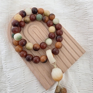 May include: A beaded necklace with a pendant and tassel, featuring a mix of brown, cream, and green beads. The necklace is displayed on a wooden tray with a carved design. The pendant and tassel add a decorative element to the jewelry.