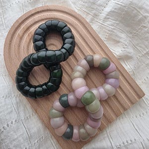 May include: Two spiral hair ties made of smooth, glossy plastic. One hair tie is solid dark forest green, while the other features a multi-colored pattern of muted pink, sage green, and beige segments arranged in a continuous circular coil design.