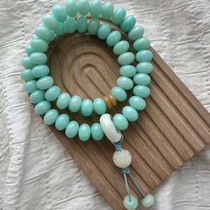 May include: A light blue beaded bracelet with a decorative tassel and a wooden display stand. The bracelet features oval-shaped beads and a white and blue tassel. The wooden stand has a natural wood grain finish.