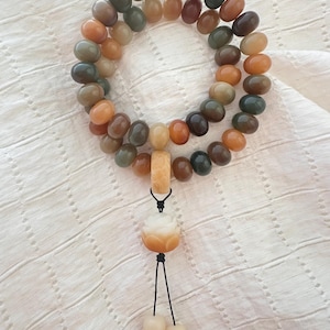 May include: A multi-colored beaded bracelet with a pendant. The beads are oval-shaped and range in color from green to brown to orange. The pendant is a carved lotus flower with a black cord and two dangling beads.