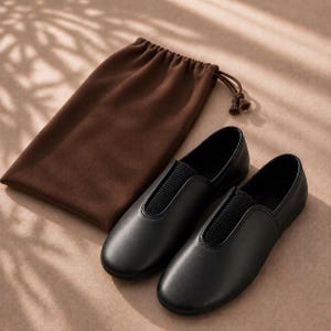 Peut inclure: Une paire de chaussures à enfiler noires avec un bout arrondi et un panneau élastique noir texturé sur le cou-de-pied, accompagnée d'une pochette à cordon en tissu marron avec un cordon assorti et deux petits pompons à l'extrémité.