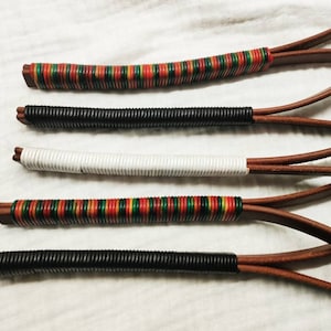 Puede incluir: Cinco correas de cuero con diferentes envolturas de colores. Las envolturas son rayas rojas, verdes y amarillas, negras, blancas y negras de nuevo. Las correas son todas de cuero marrón.