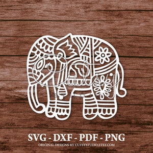 May include: A white silhouette of an elephant with intricate patterns and designs. The elephant is standing on a brown wooden background. The text "SVG - DXF - PDF - PNG" is below the elephant.