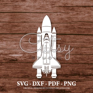 Puede incluir: Una silueta blanca de una lanzadera espacial sobre un fondo de madera marrón. La imagen incluye el texto "SVG - DXF - PDF - PNG" y "ORIGINAL DESIGNS BY CUTSYSTUDIO.ETSY.COM".