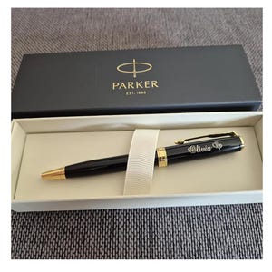 May include: A black ballpoint pen with gold-tone accents and the name Olivia engraved on the barrel, resting in a white presentation tray inside a black Parker gift box featuring the brand logo and the text EST. 1888 in gold lettering.