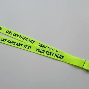 Puede incluir: Un cord&oacute;n de color amarillo ne&oacute;n con el texto negro repetido "YOUR TEXT HERE" y "ANY NAME ANY TEXT" impreso a lo largo de la cinta de tela. El cord&oacute;n incluye un cierre de seguridad de pl&aacute;stico negro y un gancho giratorio de metal plateado.