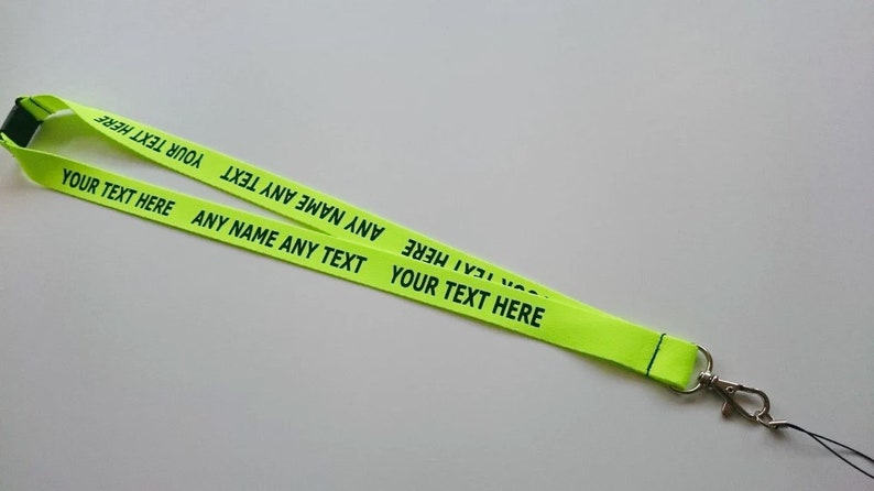 Puede incluir: Un cord&oacute;n de color amarillo ne&oacute;n con el texto negro repetido "YOUR TEXT HERE" y "ANY NAME ANY TEXT" impreso a lo largo de la cinta de tela. El cord&oacute;n incluye un cierre de seguridad de pl&aacute;stico negro y un gancho giratorio de metal plateado.