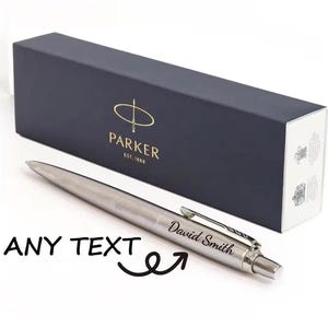 以下が含まれることがあります： 「David Smith」と刻印されたシルバーのパーカーペン。ネイビーブルーの箱にはパーカーのロゴと文字が描かれています。箱は長方形で、金色の文字と白い縁取りが施されています。