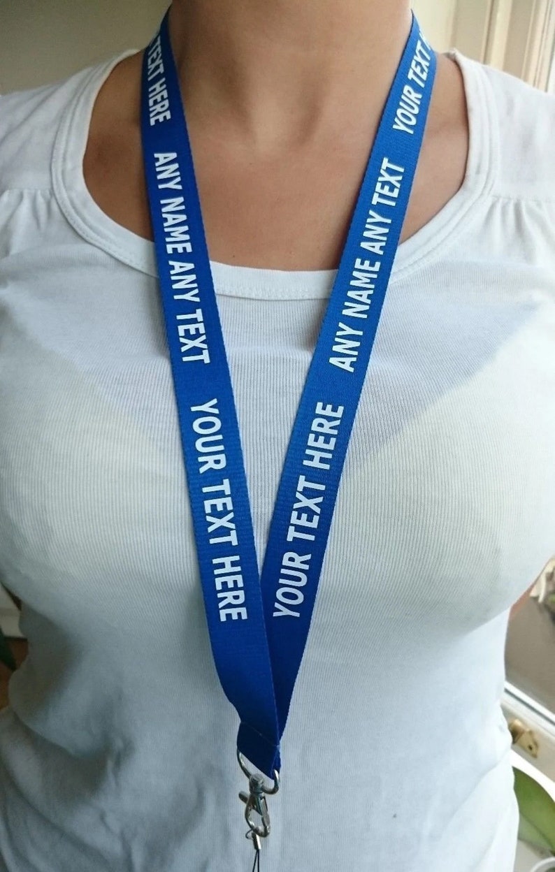 Puede incluir: Un cord&oacute;n azul de tela con el texto blanco repetido ANY NAME ANY TEXT y YOUR TEXT HERE. El cord&oacute;n cuenta con un cierre de mosquet&oacute;n de metal en la parte inferior para sujetar objetos, ofreciendo un accesorio personalizable para llevar tarjetas o llaves.