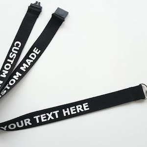 Puede incluir: Un cord&oacute;n de tela negro con texto impreso en blanco que dice CUSTOM MADE y YOUR TEXT HERE. El cord&oacute;n incluye un conector de hebilla de pl&aacute;stico negro en la parte superior y un mosquet&oacute;n de metal plateado con un lazo de cuerda negro fino.