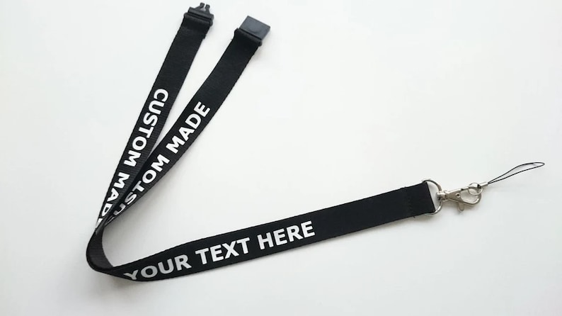Puede incluir: Un cord&oacute;n de tela negro con texto impreso en blanco que dice CUSTOM MADE y YOUR TEXT HERE. El cord&oacute;n incluye un conector de hebilla de pl&aacute;stico negro en la parte superior y un mosquet&oacute;n de metal plateado con un lazo de cuerda negro fino.