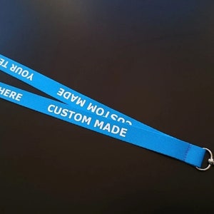 Puede incluir: Un cord&oacute;n de tela azul brillante con el texto impreso en blanco "YOUR TEXT HERE" y "CUSTOM MADE" repetido a lo largo de la cinta. El cord&oacute;n incluye un conector de seguridad de pl&aacute;stico negro en la parte superior y un cierre de mosquet&oacute;n de metal plateado.