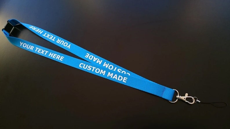 Puede incluir: Un cord&oacute;n de tela azul brillante con el texto impreso en blanco "YOUR TEXT HERE" y "CUSTOM MADE" repetido a lo largo de la cinta. El cord&oacute;n incluye un conector de seguridad de pl&aacute;stico negro en la parte superior y un cierre de mosquet&oacute;n de metal plateado.