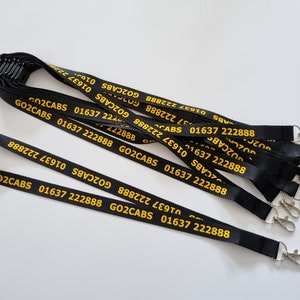 Puede incluir: Una colecci&oacute;n de cordones negros con el texto amarillo GO2CABS 01637 222888 impreso repetidamente a lo largo de la tela. Cada cord&oacute;n incluye un cierre de seguridad de pl&aacute;stico negro en la parte superior y un mosquet&oacute;n de metal plateado al final.