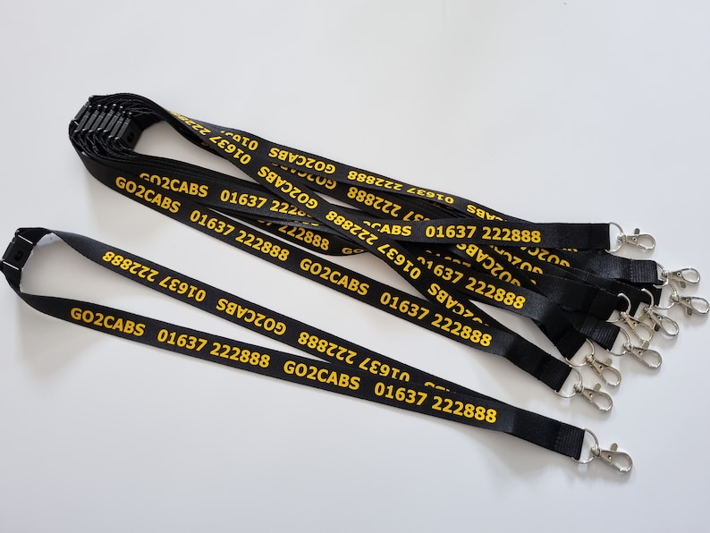 Puede incluir: Una colecci&oacute;n de cordones negros con el texto amarillo GO2CABS 01637 222888 impreso repetidamente a lo largo de la tela. Cada cord&oacute;n incluye un cierre de seguridad de pl&aacute;stico negro en la parte superior y un mosquet&oacute;n de metal plateado al final.