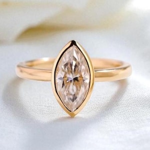May include: A gold ring with a marquise-cut diamond set in a bezel setting. The ring is displayed on a white surface, with a soft focus background. The ring's band is a simple, polished gold.