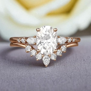 Può includere: Un set di anelli di fidanzamento in oro rosa con un grande diamante centrale a taglio ovale e una fascia abbinata. La fascia presenta diamanti più piccoli, creando un design scintillante ed elegante. L'anello è su uno sfondo grigio chiaro.