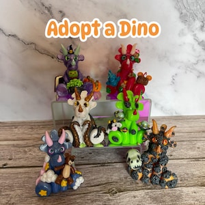 Peut inclure: Adoptez un Dino ! Six figurines de dinosaures en pâte polymère faites à la main dans différentes couleurs et designs. Les dinosaures sont assis sur une surface en bois.
