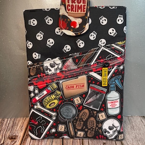 May include: Black fabric case with a flap closure, featuring a repeating skull pattern. The front has a zippered pocket with crime scene-themed graphics, including a magnifying glass, evidence bag, and the text "CRIME SCENE". The flap reads "TRUE CRIME".