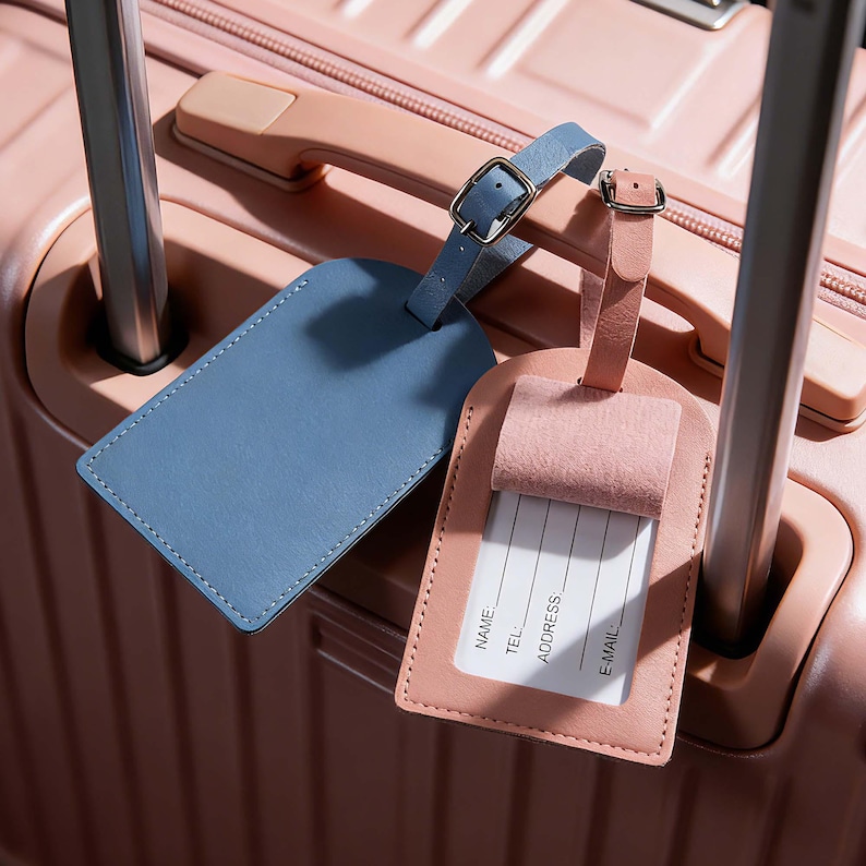 May include: Two rectangular luggage tags with adjustable buckle straps. One tag is solid blue, and the other is light pink with a flap covering a white identification card featuring the printed fields Name, Tel, Address, and E-mail.
