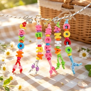 Op de afbeelding: Vijf gepersonaliseerde sleutelhangers hangend aan een wit koord, elk voorzien van kleurrijke plastic letterkralen die namen spellen zoals JAXON, HENRY, MOBE, ANMA en COCO, geaccentueerd met kleine stervormige kralen en goudkleurige metalen sluitingen.