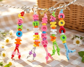 May include: Five personalized keychains hanging from a white cord, each featuring colorful plastic letter beads spelling names like JAXON, HENRY, MOBE, ANMA, and COCO, accented with small star-shaped beads and gold-toned metal flower clasps.