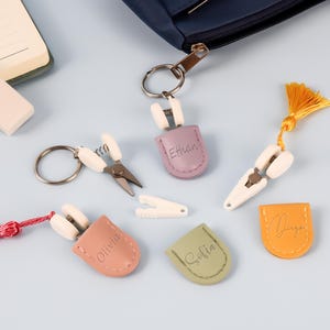 Può includere: Una collezione di portachiavi e accessori personalizzati, tra cui forbici, mollette e targhette in pelle con nomi. Gli articoli sono in vari colori come viola, arancione e rosa, con uno sfondo blu.