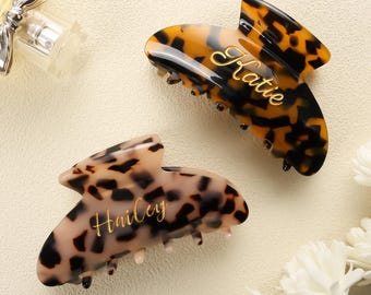 May include: Two hair claw clips with a tortoiseshell pattern. One clip is dark brown and black with the name Katie in gold script. The other clip is a lighter beige and brown with the name Hailey in gold script. Both clips feature interlocking teeth.