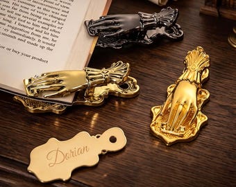 May include: A collection of metal hand-shaped page holders in gold and black finishes. The clips feature a detailed hand design with a ruffled cuff. One gold-toned metal tag is engraved with the name Dorian in a cursive script font.