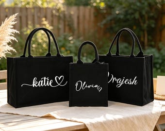 May include: Three black canvas tote bags of varying sizes, each featuring white custom text. The left bag displays the name Katie with a heart, the center bag shows Olivia with a heart, and the right bag features the name Rajesh in white script.