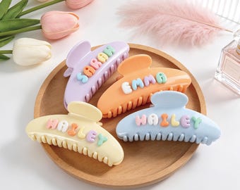 May include: Four pastel-colored hair claw clips with rounded teeth. The lavender clip features the name SARAH, the orange clip features EMMA, and two light yellow and light blue clips feature the name HAILEY in raised, multi-colored bubble letters.