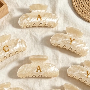 May include: A collection of cream-coloured hair claw clips with a marbled finish. Each clip features a single gold-toned letter, including A, C, and Y, centred on the front. The clips have a curved, multi-pronged design for securing hair.