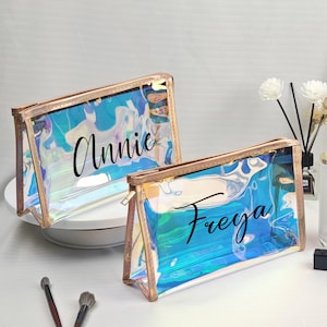 May include: Two iridescent cosmetic bags with gold-colored trim. One bag has the name "Annie" and the other has the name "Freya" in black script. The bags are clear, allowing the contents to be visible.