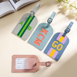 May include: Three rectangular luggage tags with adjustable straps and buckles, featuring colorful stripes and letters A, B, KCM, and GO. A fourth pink luggage tag shows a white identification card slot with fields for name, telephone, address, and email.