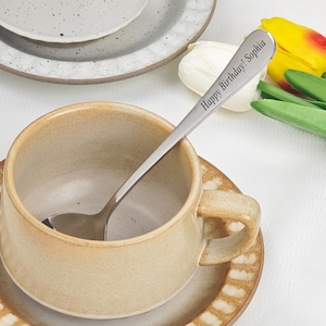 Può includere: Un cucchiaio in acciaio inossidabile con la scritta "Happy Birthday! Sophia" incisa poggia in una tazza di ceramica beige su un piattino abbinato. Un secondo piatto e dei fiori sono sullo sfondo. Il cucchiaio è per servire.