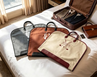 May include: Three garment bags in gray, brown, and cream, each featuring dual handles and personalized embroidery. The cream bag displays the name JACKSON with crossed golf clubs. A brown suitcase containing folded clothing and a small brown briefcase are nearby.