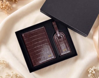 May include: A dark brown, crocodile-embossed passport holder and luggage tag set presented in a black gift box. Both items feature the gold-toned initials AH embossed on the front. The set is arranged neatly within a black foam insert.