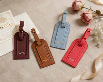 May include: Four rectangular luggage tags with rounded tops and adjustable buckle straps. The tags are dark burgundy with WYA, tan with DNY, light blue with ESP, and red with QIM embossed on them. Each tag features a stitched arch design.