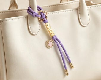 May include: A cream-colored handbag featuring a purple cord accessory attached to the handle. The accessory includes a gold-toned flower charm, gold cube beads spelling the name JULIE, and a circular pendant with a pink flower and crystals.