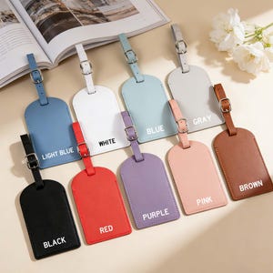 May include: A collection of nine rectangular luggage tags with rounded bottoms, each featuring a matching strap and a silver-toned metal buckle. The tags are available in black, light blue, white, blue, gray, red, purple, pink, and brown colors.