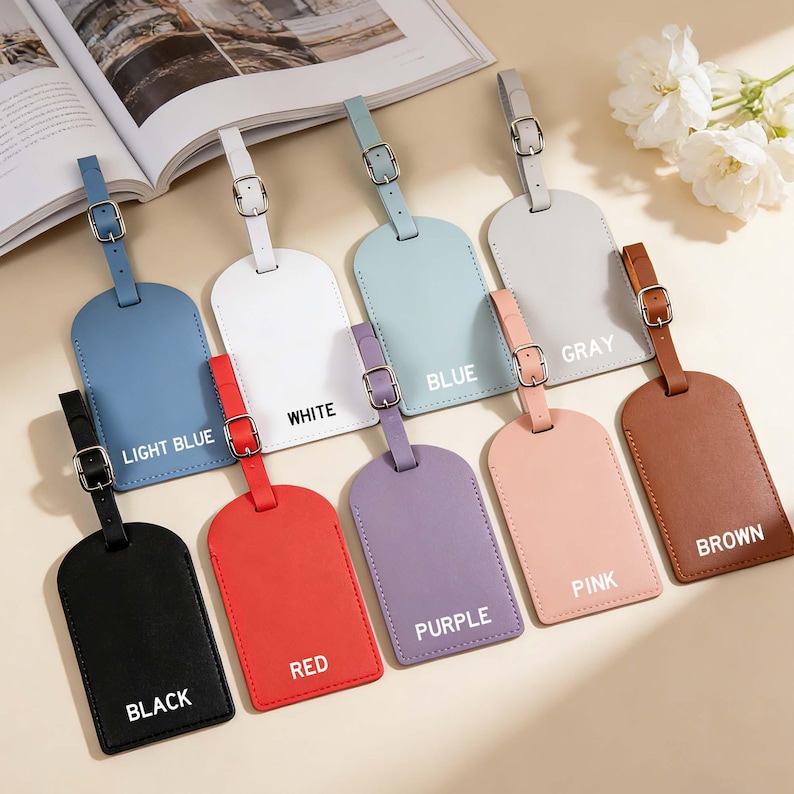 May include: A collection of nine rectangular luggage tags with rounded bottoms, each featuring a matching strap and a silver-toned metal buckle. The tags are available in black, light blue, white, blue, gray, red, purple, pink, and brown colors.