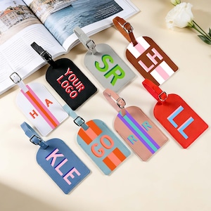 May include: A collection of eight rectangular luggage tags in various colours including black, white, grey, brown, blue, and red. Each tag features a buckle strap and personalised lettering or text such as YOUR LOGO, SR, LH, HA, GO, RR, LL, and KLE.