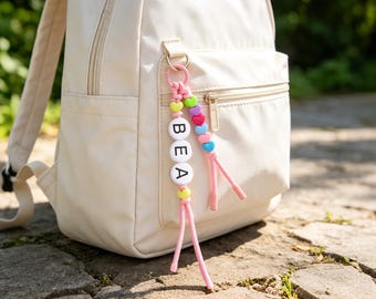 May include: A personalized keychain attached to a cream-colored backpack. The keychain features white circular beads with the black letters B, E, and A, accented by smaller heart-shaped beads in pink, purple, blue, yellow, and green on pink cords.