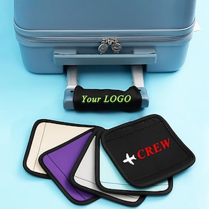 Op de afbeelding: Een blauwe koffer met een zwarte handgreep met de groene tekst "Your LOGO". Hieronder worden verschillende handgreepbeschermers in verschillende kleuren getoond, waaronder een zwarte met "+CREW" en een vliegtuigafbeelding.