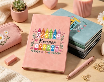 May include: A pink notebook featuring a graphic of colorful crayons and the name Bonnie, alongside a stack of notebooks in blue, green, gray, and black, a pink pencil case with embroidered daisies, a pink pen, and gold metal binder clips.