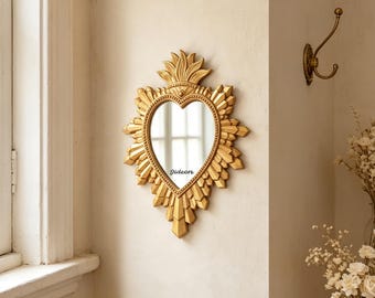 May include: A heart-shaped wall mirror with a gold-colored frame featuring a sunburst design and a flame-like crest at the top. The word Gideon is printed in black on the lower portion of the mirror glass, which reflects light from a nearby window.